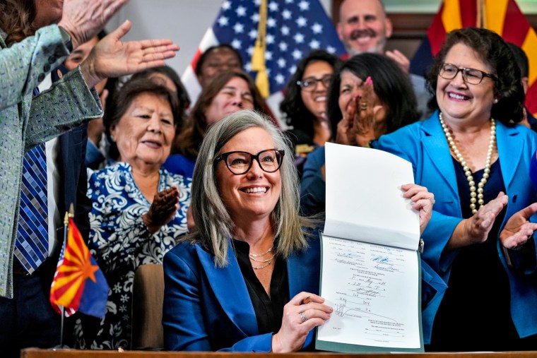 Katie Hobbs holds up a document while seated at a desk surrounded by clapping people