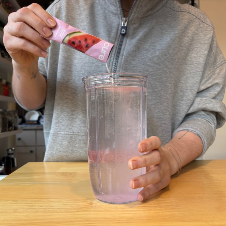 A hand pouring a single-serve packet of Cure electrolyte powder into a cup of water.