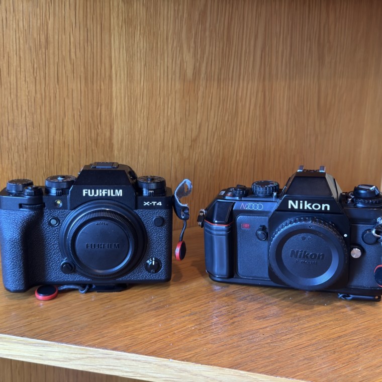 A close-up shot of the front of the Fujifilm X-T4 digital camera and the Nikon N2000 film camera on a wooden shelf.