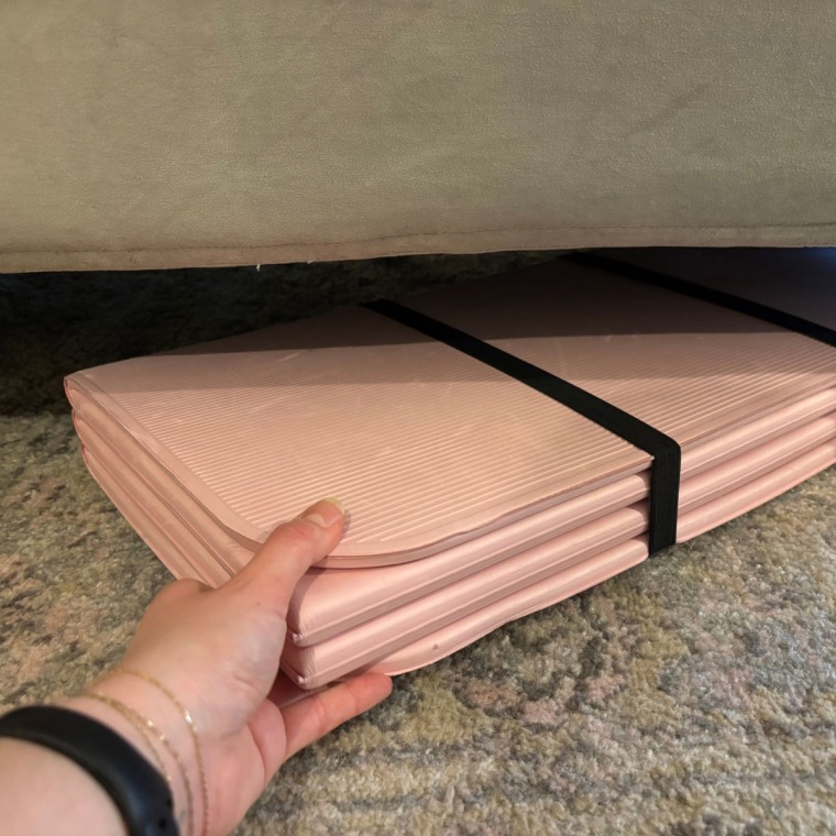 A woman’s hand holding a folded pink Stakt Mat and sliding it under a grey couch.