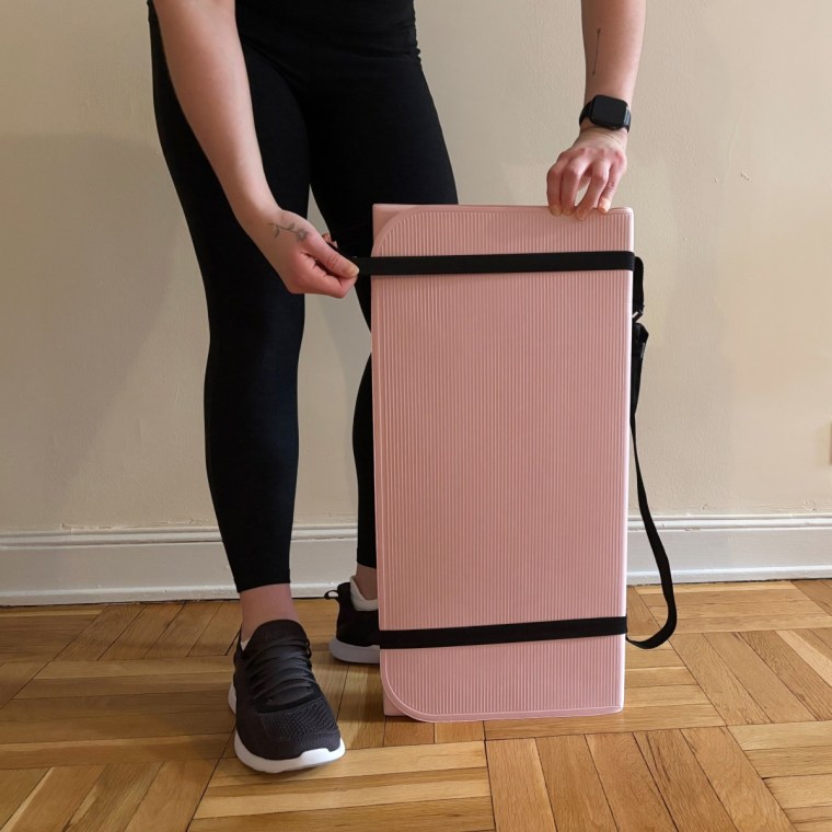 A woman putting a black strap around a pink Stakt Mat while standing in front of a white wall.