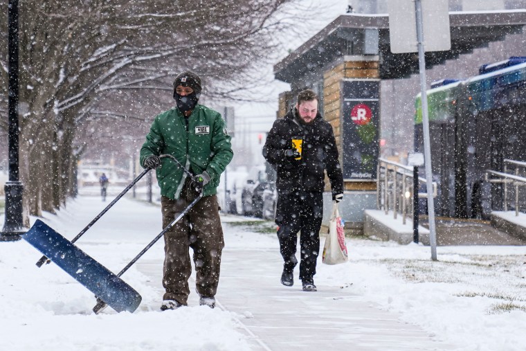 Miguel Martinez scoops snow off the sidewalk, someone walks next to him holding a drink