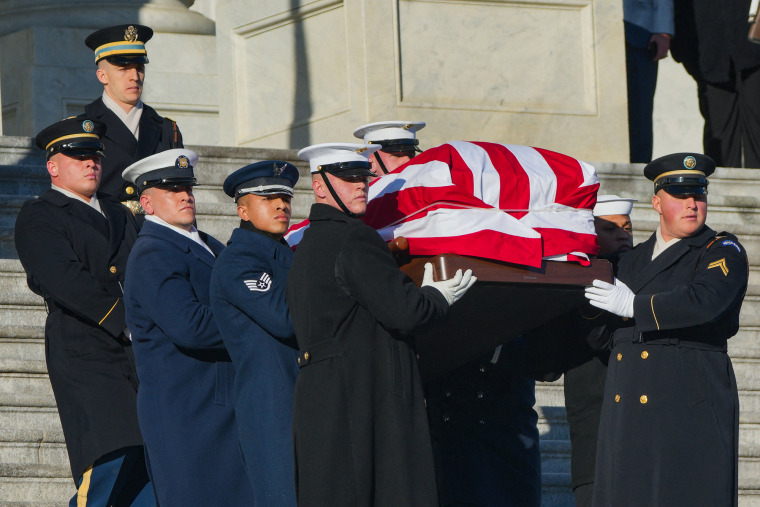 A coffin draped in an American flag is carried by several people down the stairs outside.