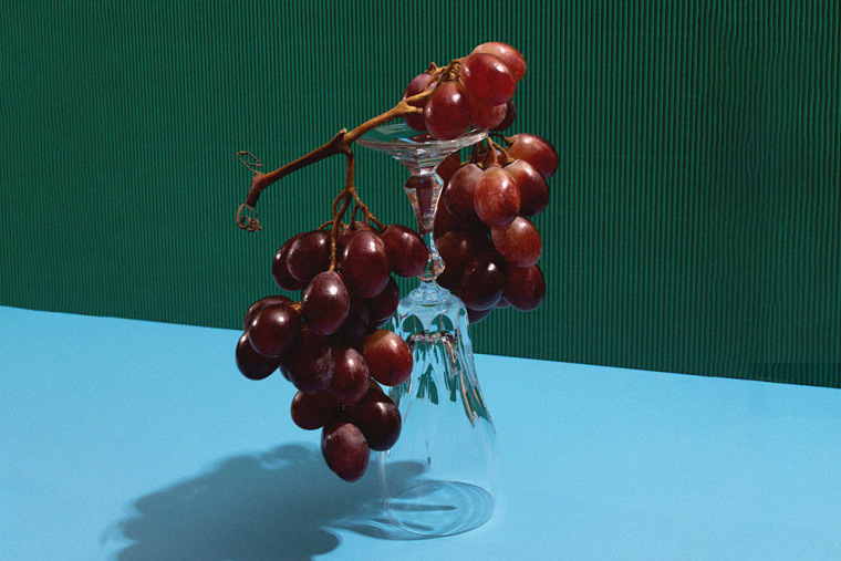 Still-life style photo of red grapes hanging over an overturned wine glass against a green and blue background 