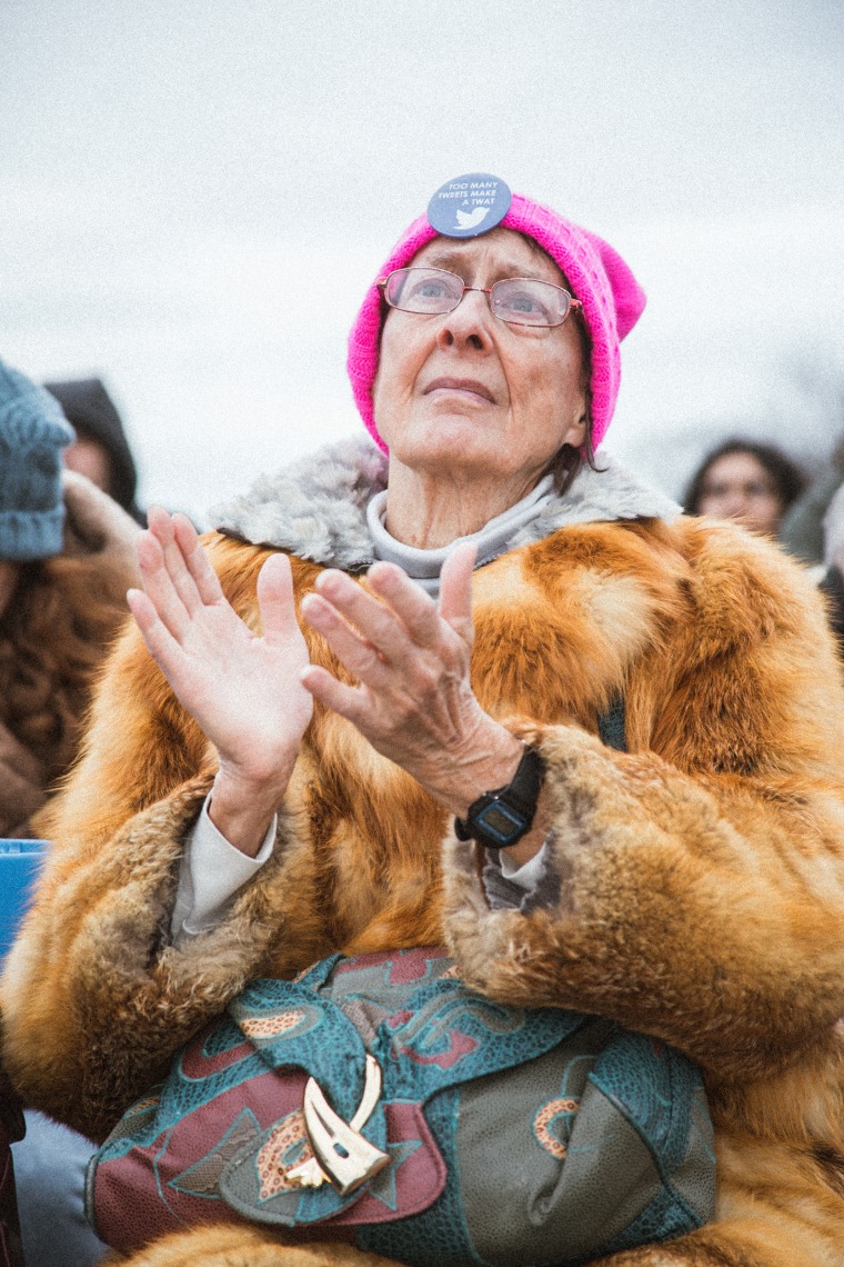 Mulher idosa com chapéu rosa olhando para cima e batendo palmas.