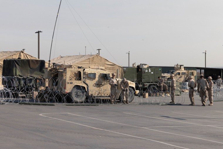 Soldiers stand outside in a parking lot with tents, barbed wire, and vehicles