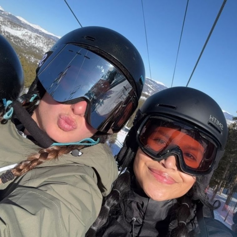 Four women wearing ski goggles standing on a ski slope against a blue sky with trees in the background.