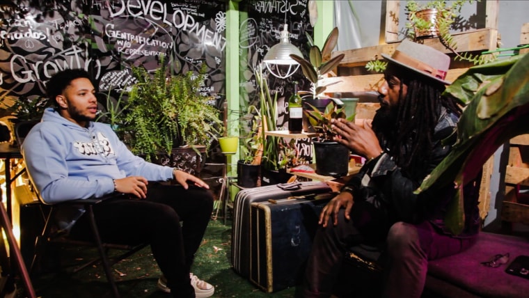 Two men speak to each other while seated inside of a room with various plants