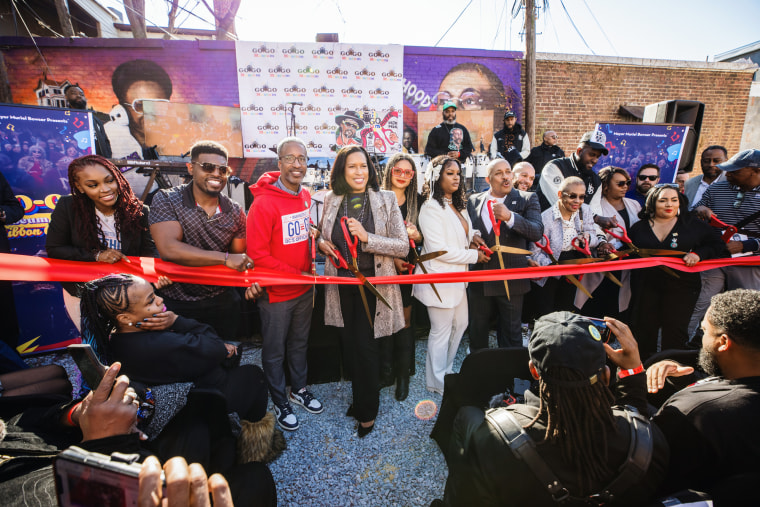 A row of people line up behind a red ribbon to cut it