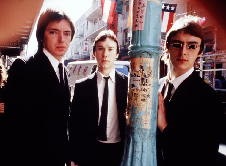 From left, Bruce Foxton, Rick Buckler, and Paul Weller wear black suits and ties while posing for a photo outside around a utility pole