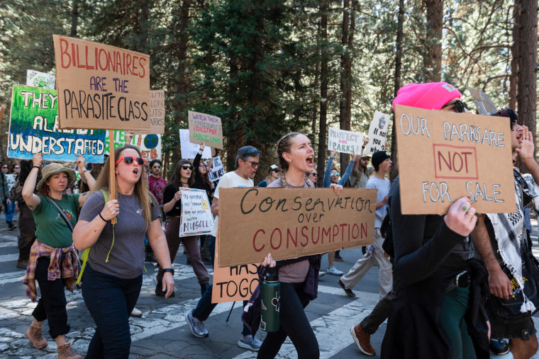 People hold signs in protest while walking on a road near a forest outside 