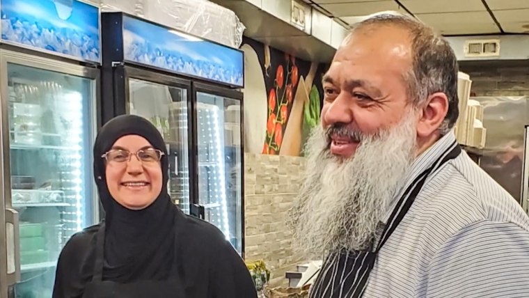 From left, Emine Emanet and Celal Emanet stand and smile in the aisle of a restaurant next to a fridge