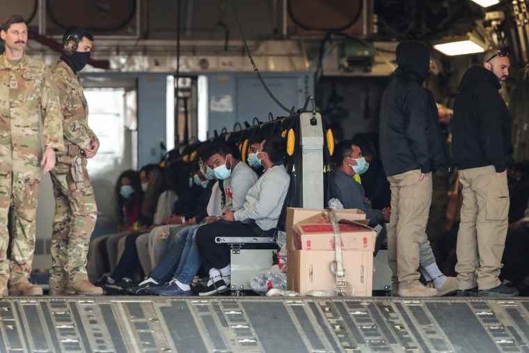 People wearing medical masks sit on a plane surrounded by soldiers and other personnel