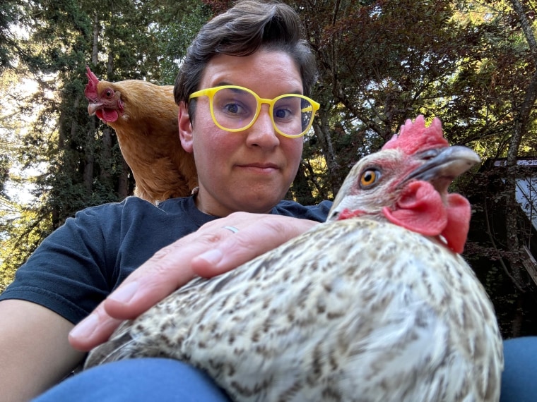 Christin New holds a chicken while another one stands on her shoulder, she wears yellow rimmed glasses