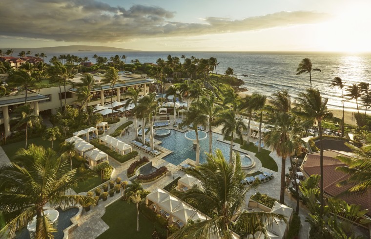 A aerial view of a hotel resort, palm trees, cabanas, a pool, and the ocean appears