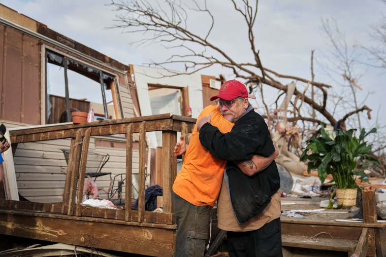 Two people hug in front of the remnants of a storm-damaged house outside