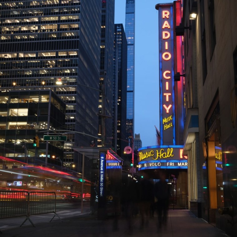 A photograph of the neon Radio City Music Hall sign at dusk. Pedestrians and traffic appear blurry and in-motion.