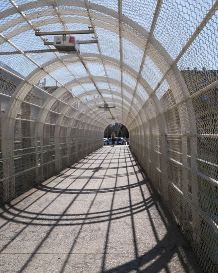 A POV photo crossing a pedestrian bridge that is surrounded by metal fencing.