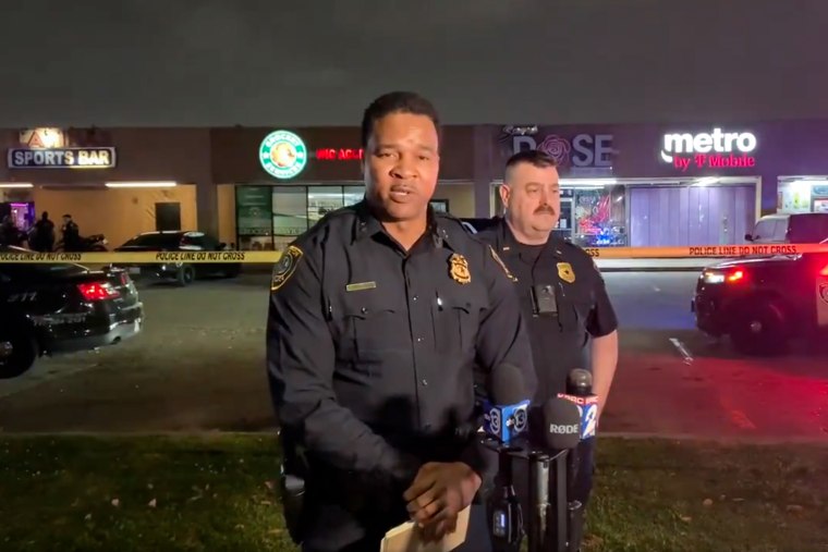 J. Skelton speaks into microphones in a parking lot with police cars and police tape, in front of another officer