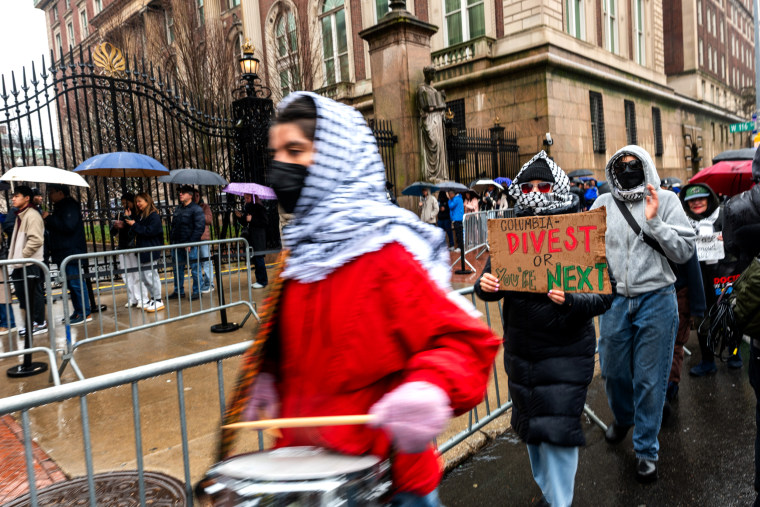 Demonstrators hold signs and bang on drums.
