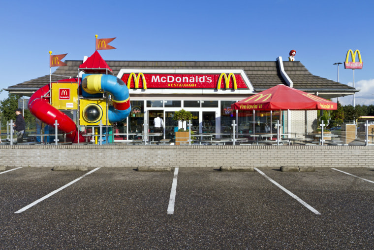 "Best, the Netherlands - September 19, 2012: Exterior view of a McDonald's restaurant with McDonald's logo on the roof and red logo advertising sign placed on a pole. McDonald's is the world's largest chain of hamburger fast food restaurants, with over 30,000 restaurants worldwide. Outside one of the employees is cleaning the tables."