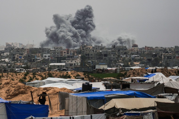 Smoke rises to the sky in the distance behind buildings and tents