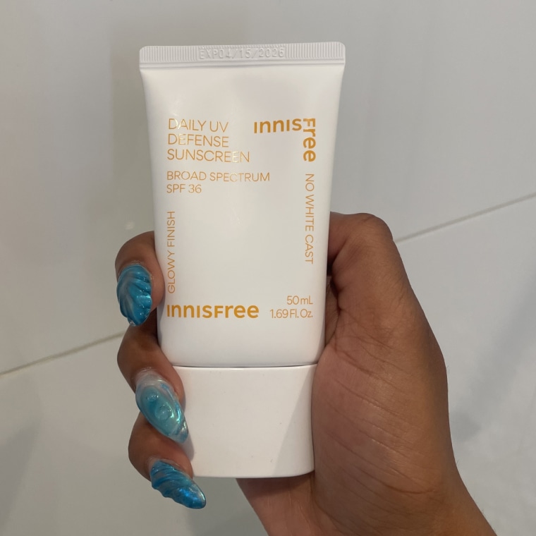 Innisfree Daily UV Defense Sunscreen being held by a hand with blue nails in front of a white tile wall.