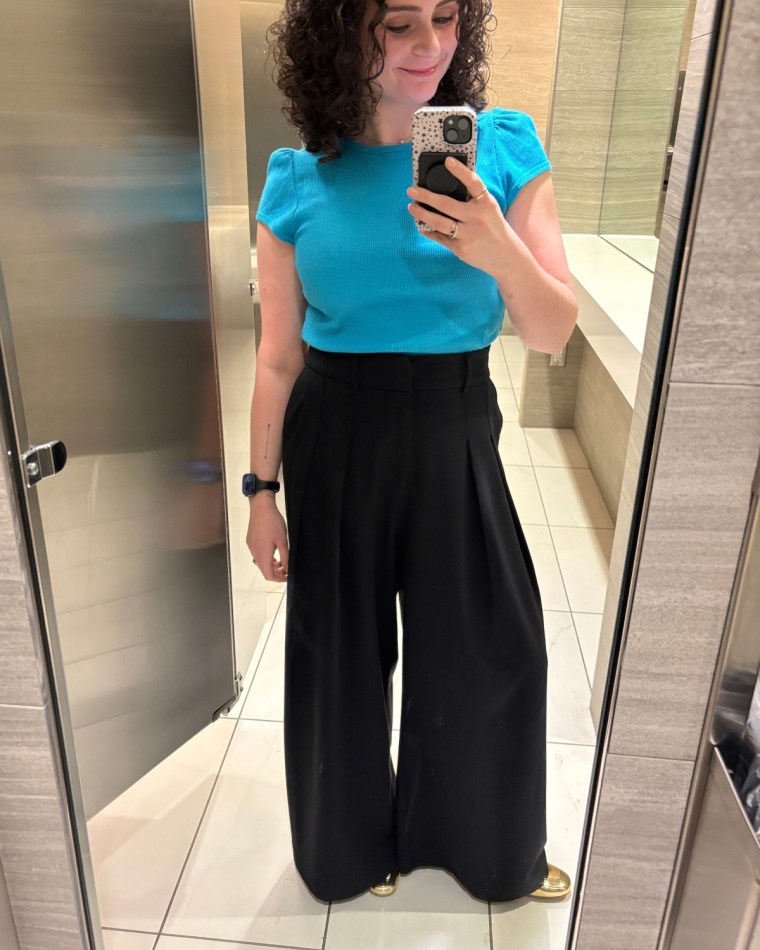 A woman taking a photo of herself in a mirror while wearing a blue shirt and black Universal Standard’s Stephanie Wide Leg Ponte Pants.