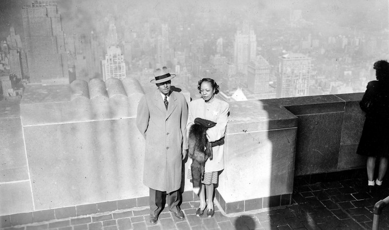 A couple on the top of the Empire State Building, on an observation deck, viewed from above, the skyline of New York visible in the background, the man wearing a trench coat and formal hat, the woman wearing a shirt and holding a fur coat,