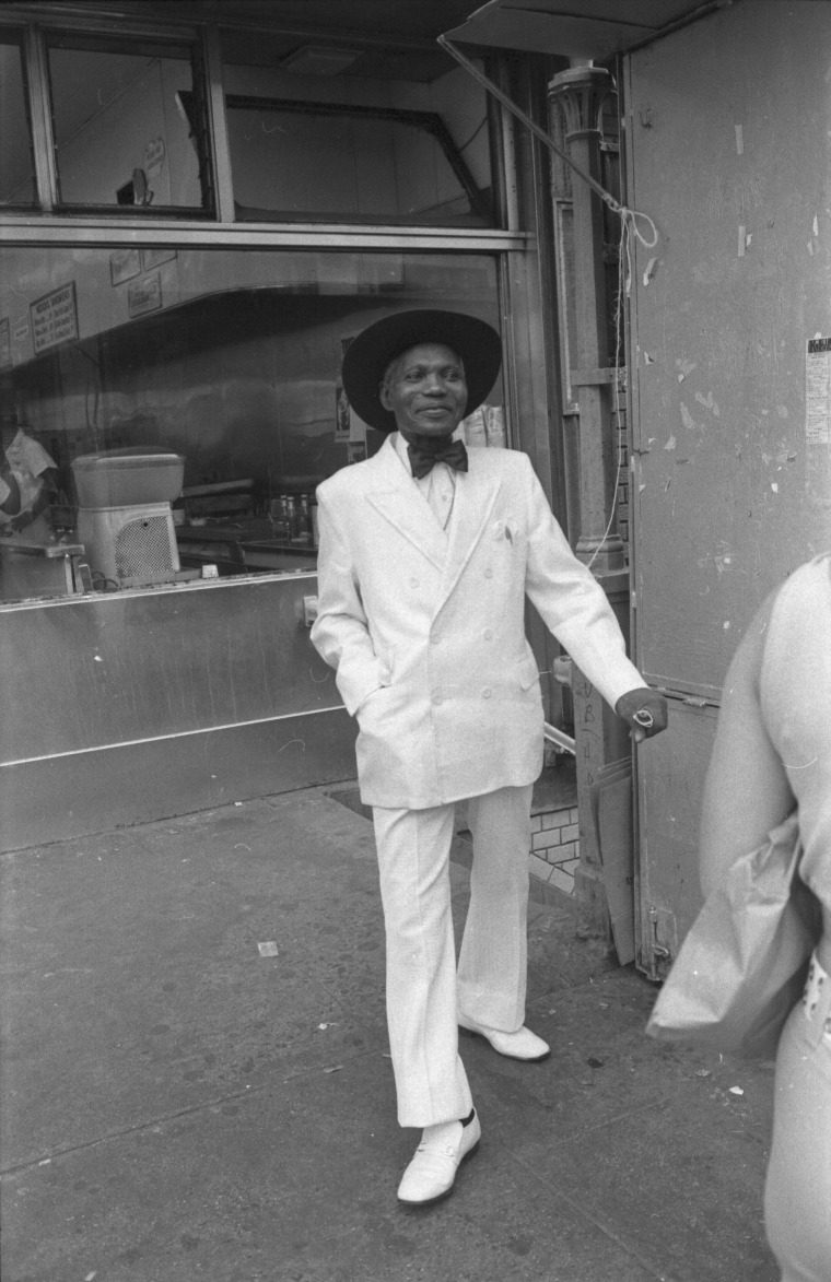 A well-dressed elderly man, wearing a white double breasted suit, bow tie, hat, and white leather shoes, smiles as he walks along an unidentified Harlem street, New York, late twentieth century.