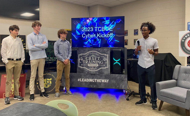 Four people stand near a tv screen in a class room