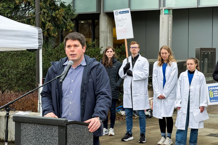 Nick Chavkin, an assistant professor in the pediatrics department at UW Medicine, speaks out against cuts to NIH funding at a Feb. 19 rally in Seattle. Castelli, a doctoral student at UW, holds a sign decrying the cuts in the background.