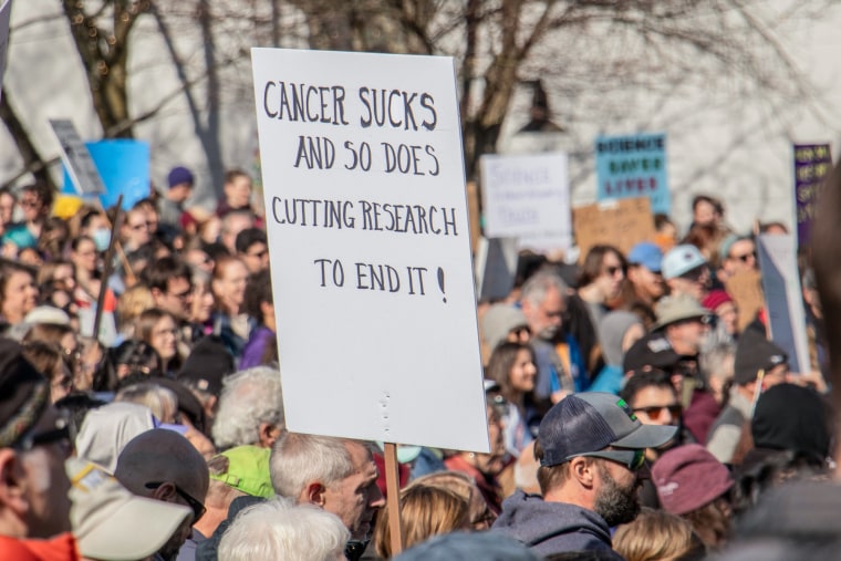Above a crowd of people, a sign is held that reads "Cancer sucks, and so does cutting research to end it!"