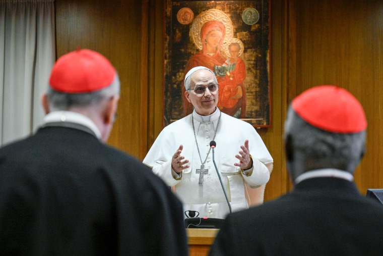 Pope Leo speaks and gestures his hands as two people watch