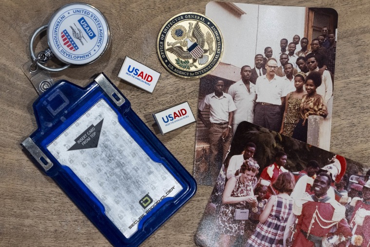 Anna Eisenberg's empty USAID badge holder is seen next to USAID pins, a coin from the USAID Office of Inspector General awarded to her mother, Cathy Votaw, and photographs of her grandfather, Albert Votaw, and his family during his service with USAID in West Africa