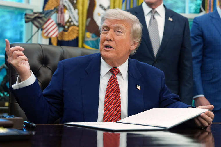 Donald Trump speaks while gesturing and holding a document at his desk