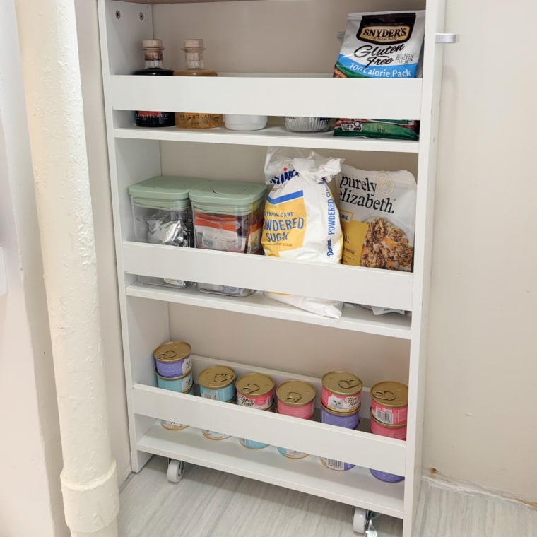 A white, three-shelf rolling cart with a wood top against a white wall in a kitchen.