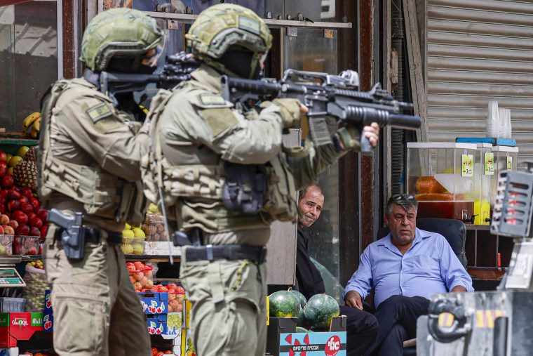 Israeli army soldiers aim their rifles as Palestinians sitting outside a shop look on 