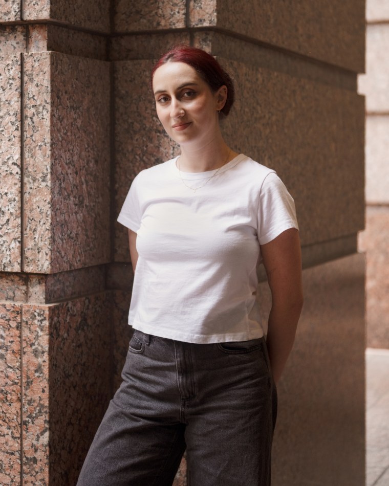 A model leans against a stone pillar while wearing a white t-shirt and black barrel leg jeans.