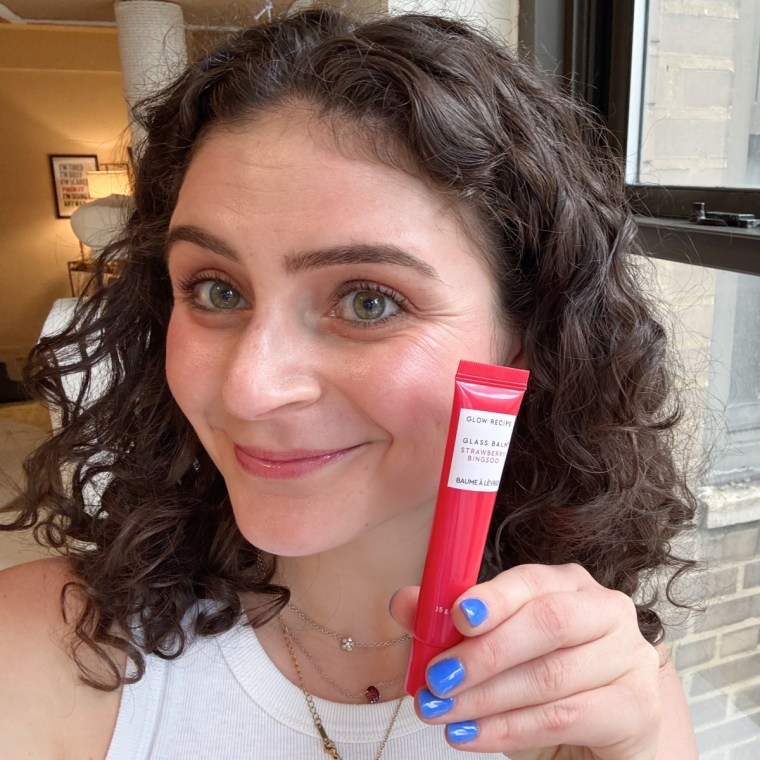 A woman holding a tube of Glow Recipe’s Glass Balm next to her face while wearing the balm on her lips.