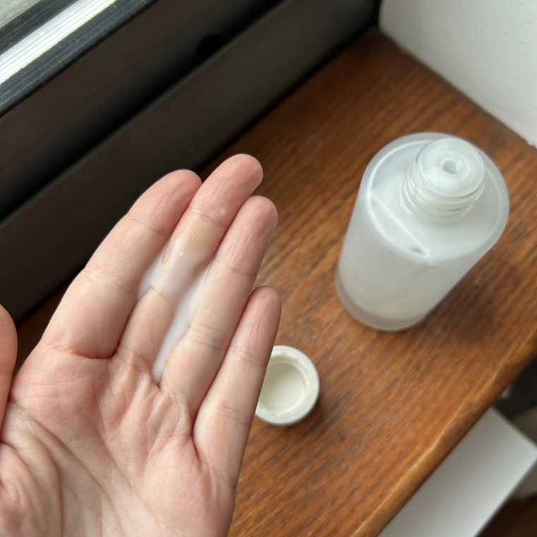 A hand holding a small pump of Ilia The Base Face Milk in beside the Ilia bottle on a windowsill.