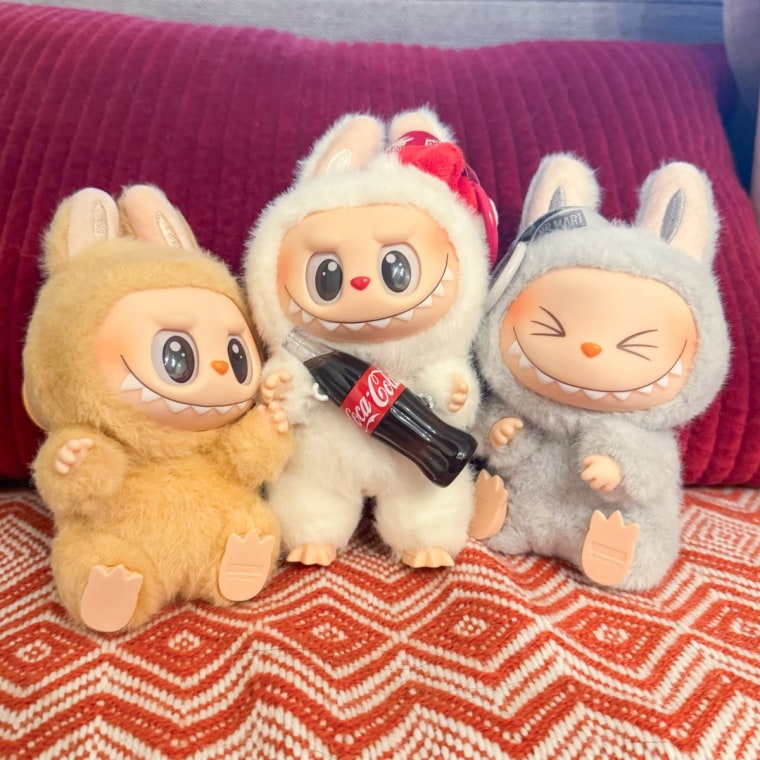 Three Labubu’s sitting side by side on a blanket covered bench. From left to right: Grey-blue Labubu with squinted eyes, white Labubu with Santa hat on right ear and glass Coca-Cola bottle in hands, light brown Labubu sitting down