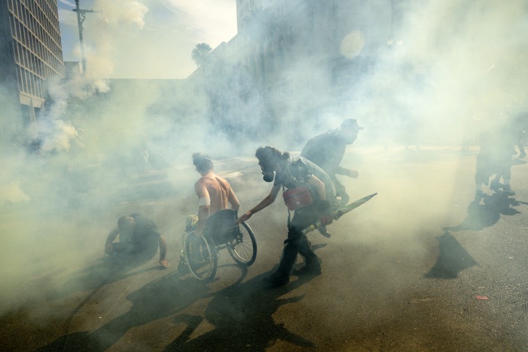 A man in a wheelchair and others move through tear gas clouds.