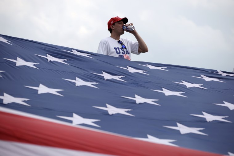 Image: A man drinks a beverage while holding a giant American flag