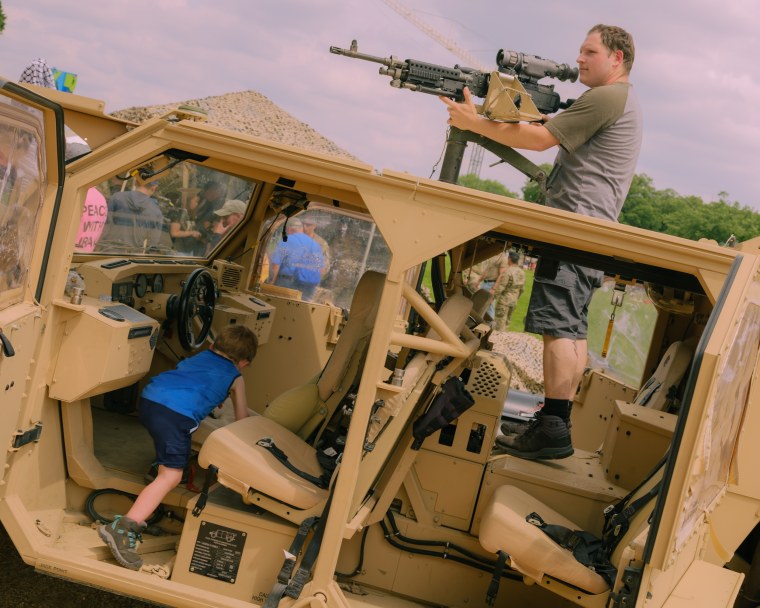 A man poses with a large gun atop a military vehicle, while a young boy climbs on the front seats.