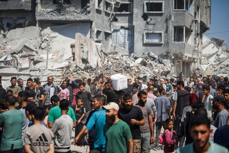A large group of people walk outside, in the center a person holds a white box on their shoulder, rubble of a building in the background