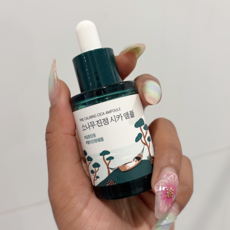 A left hand with multicolored nails holding the Round Lab Pine Calming Cica Ampoule bottle in front of a white tiled bathroom wall.