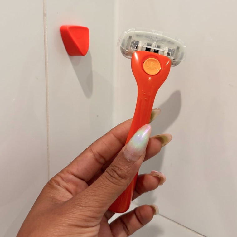 A left hand holding the Billie razor beside the magnetic holder in front of a tiled shower wall.