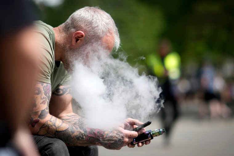 A man vapes while sitting and looking at his cell phone.