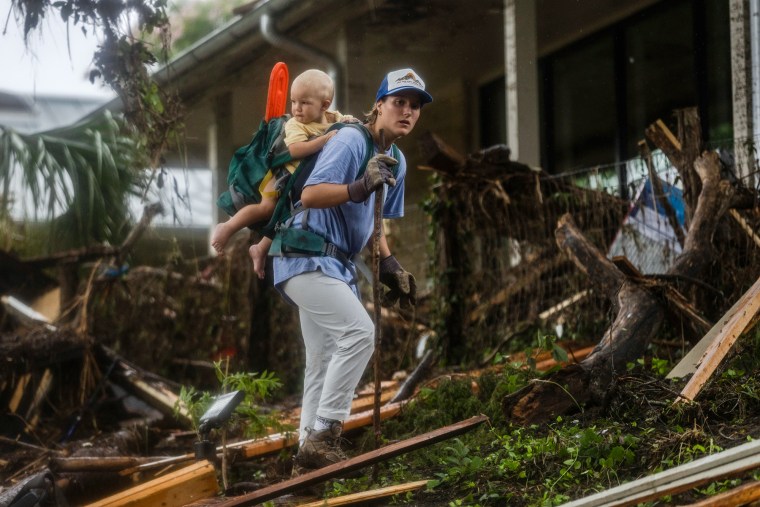 A person walks with a waking stick carrying a baby on their back outside through debris